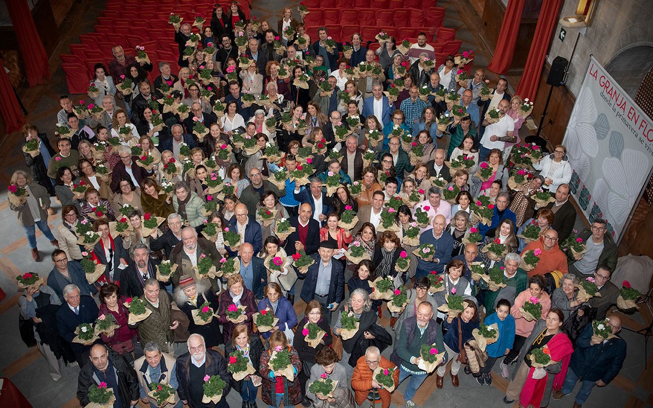 A large group of event attendees poses for an aerial photo holding pink flower bouquets indoors.
