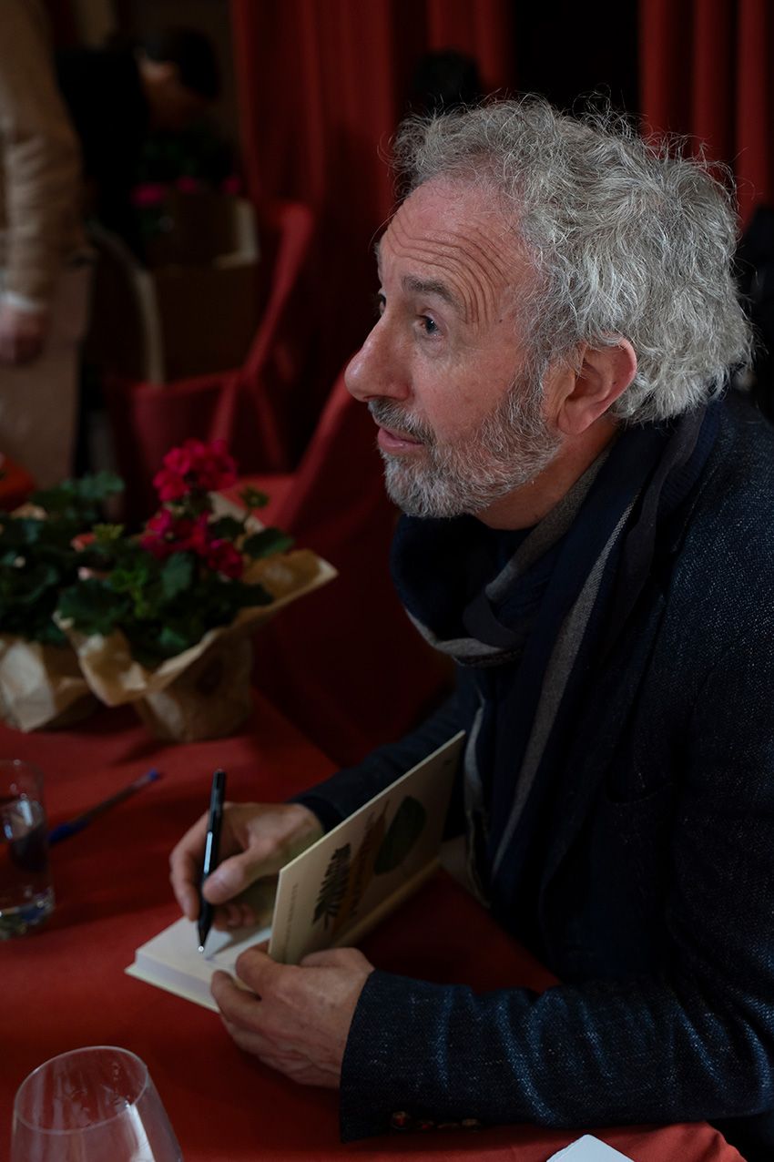 Curly-haired man with beard signing autographs at table with red floral arrangements nearby.