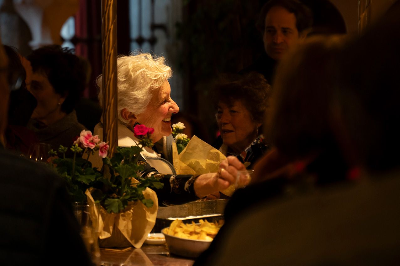 Elderly white-haired woman smiling at lively indoor gathering with pink floral arrangements.