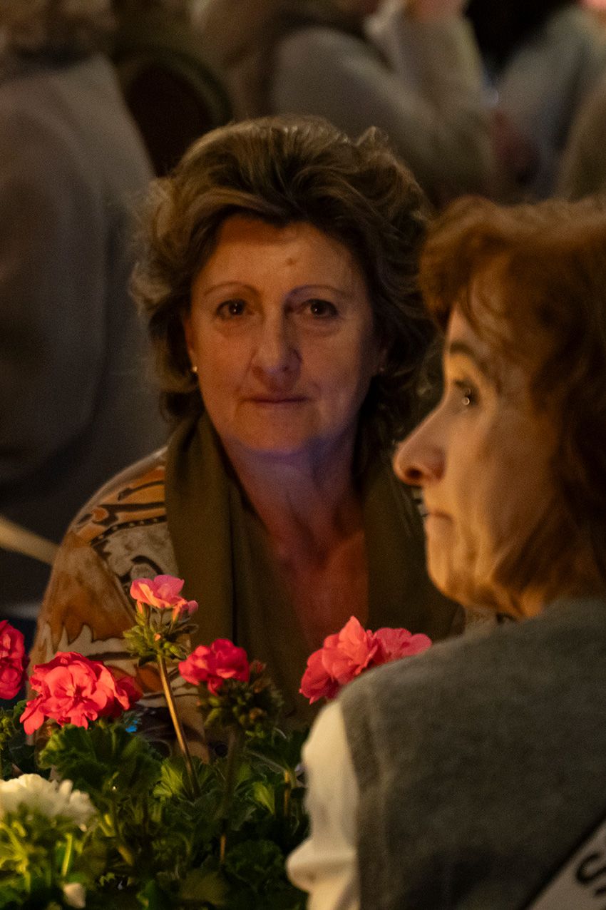 Two women in conversation at an event, with vibrant pink geraniums in the foreground.