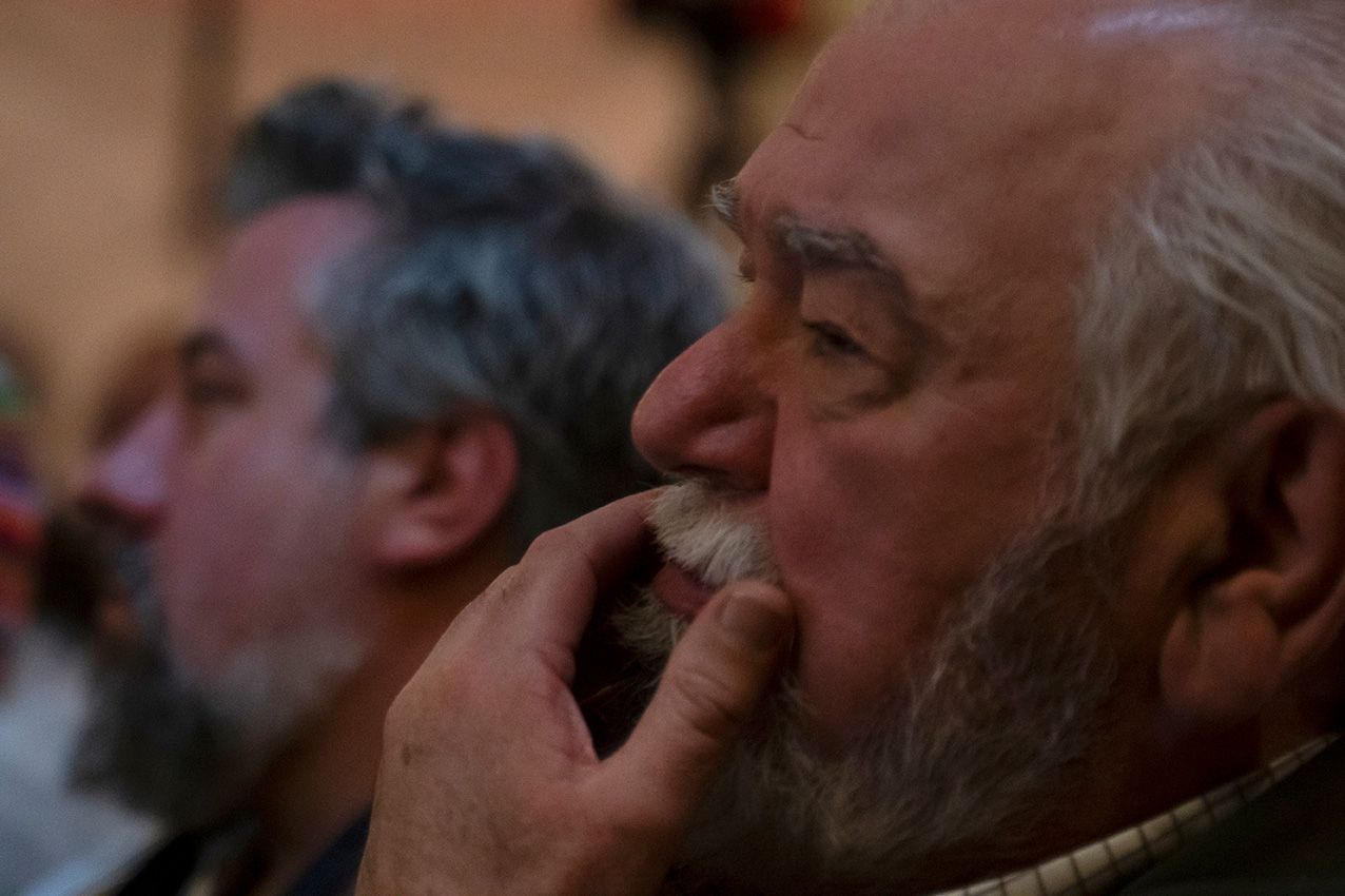 Elderly bearded man listens thoughtfully during a conference presentation in a dimly lit venue.
