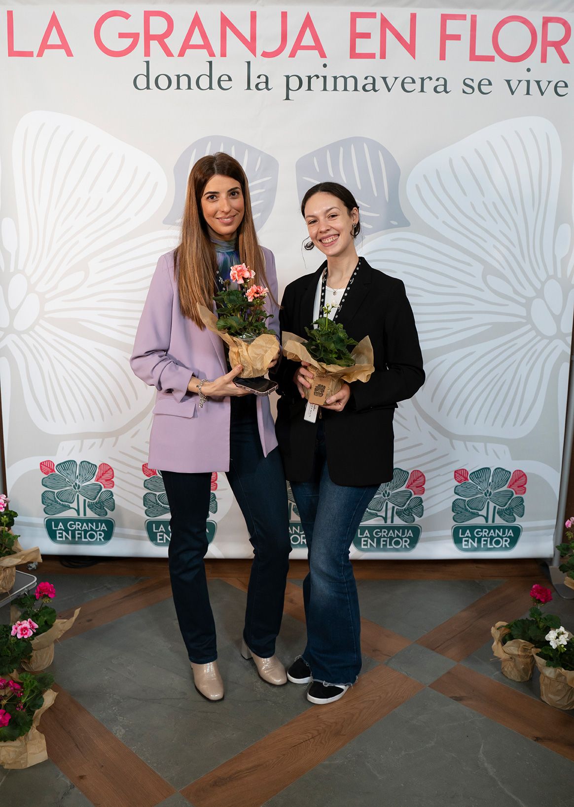Two women holding potted flowers at La Granja en Flor spring event, one in lavender blazer.