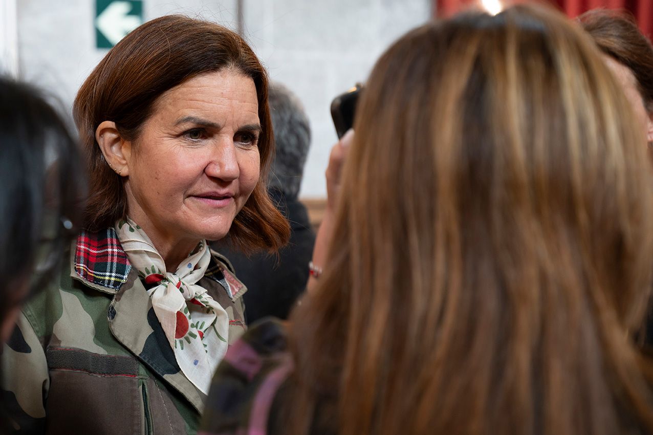 Woman in military green jacket wearing plaid scarf conversing at an indoor social event.