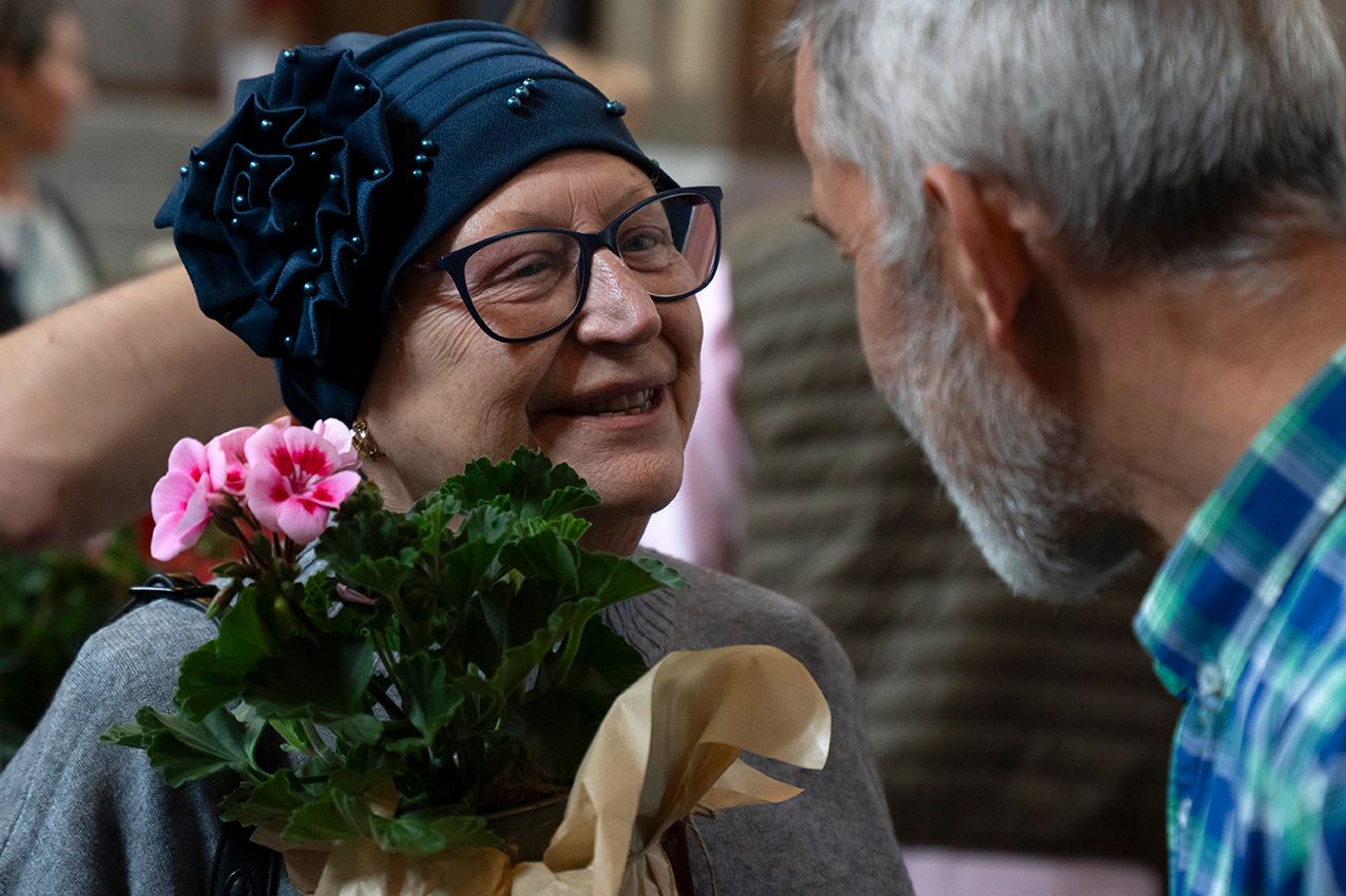 Smiling woman wearing blue headscarf and glasses holds pink geranium plant at spring flower event.