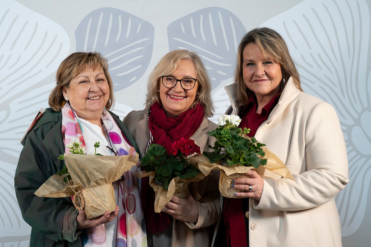 Three women smiling and holding flower bouquets wrapped in brown paper at an event.