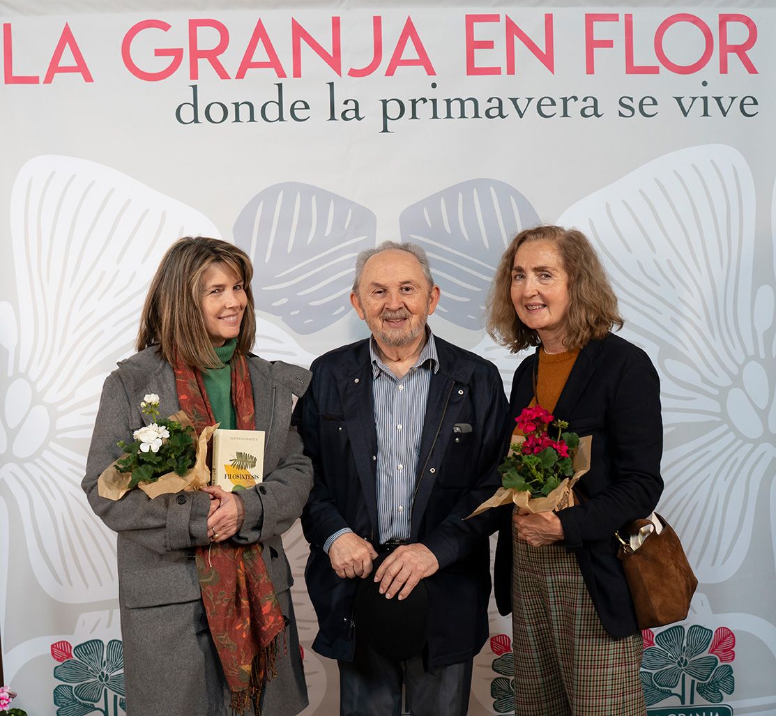 Three people smile posing in front of La Granja en Flor spring flower festival banner holding plants.
