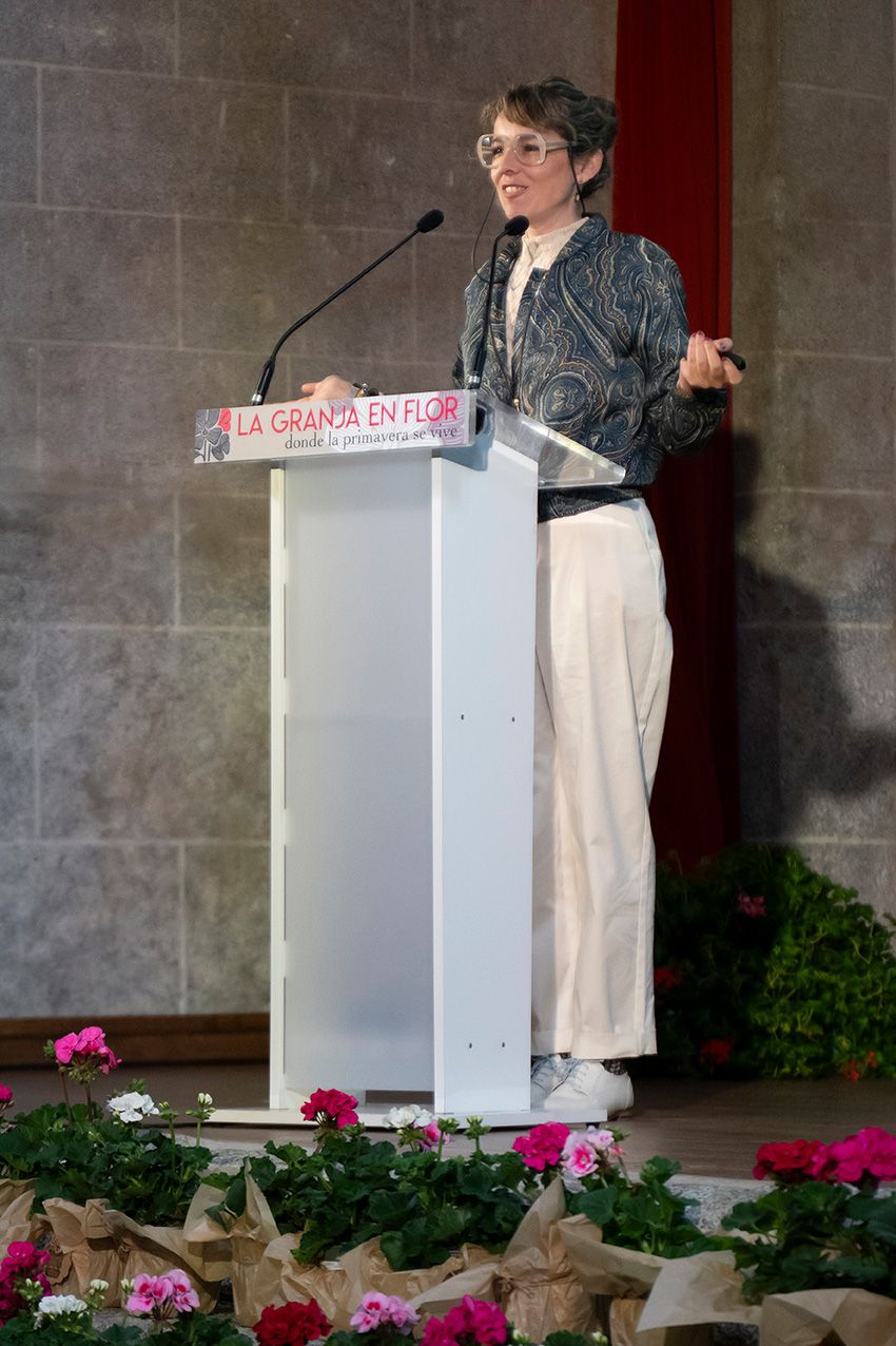 Woman speaking at podium at La Granja en Flor spring festival with flowers nearby.