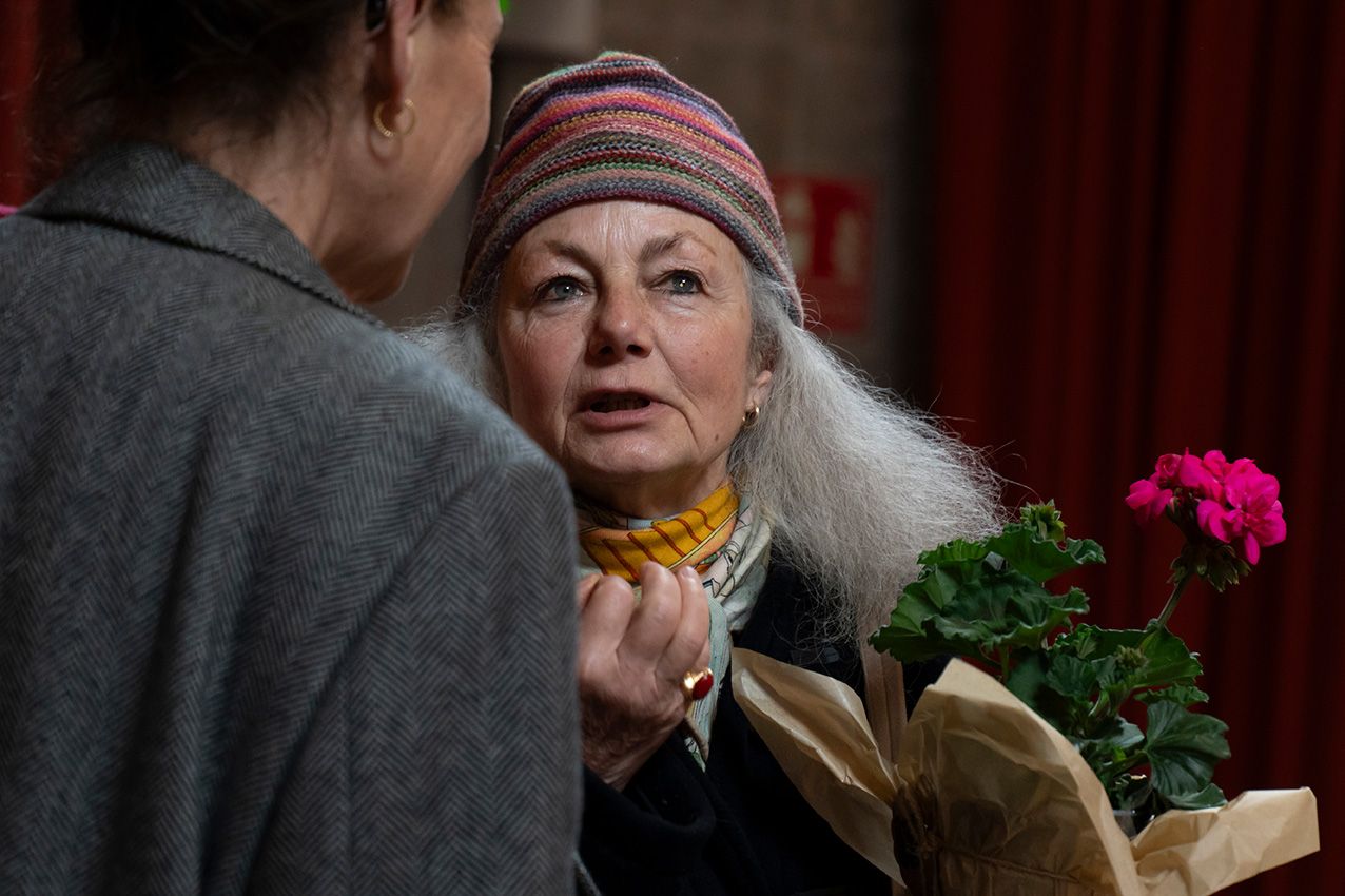 Older woman with long gray hair and striped hat holding pink geranium while talking at indoor event.