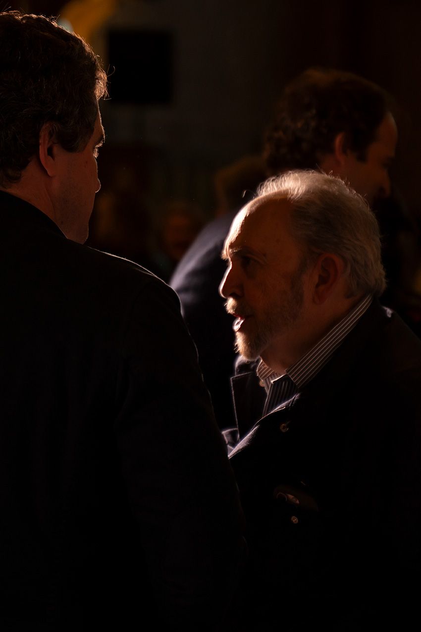 Two men in dark suits engaged in conversation at a dimly lit evening event.