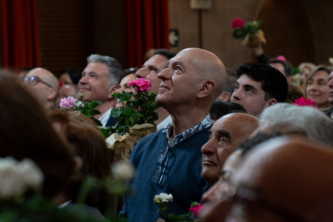 Bald man holding pink flowers stands among a large crowd at an indoor floral distribution event.