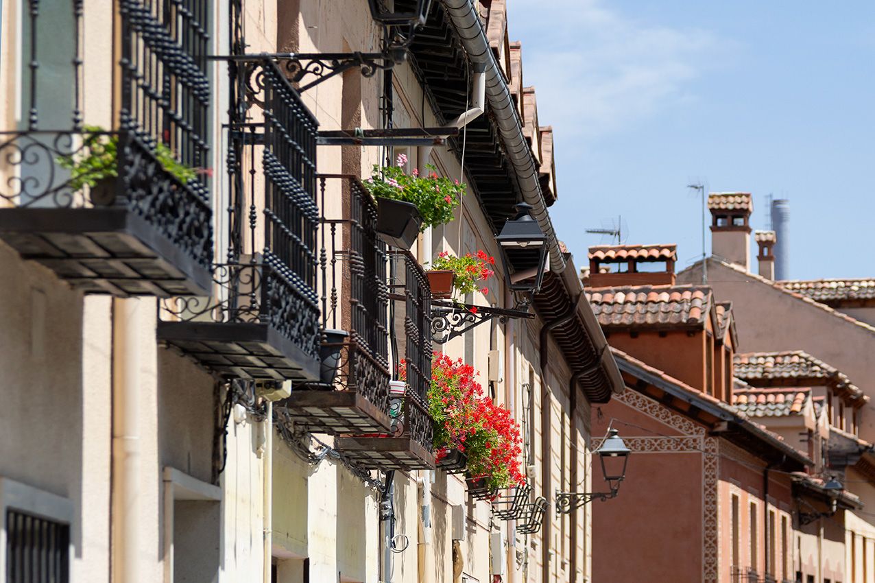 Historic European street with ornate iron balconies adorned with vibrant red geraniums on a sunny day.