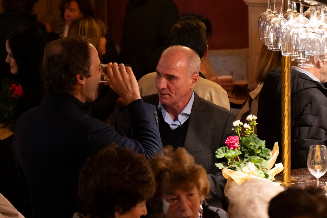 Two men in deep conversation at crowded indoor event with pink flower centerpiece on table.