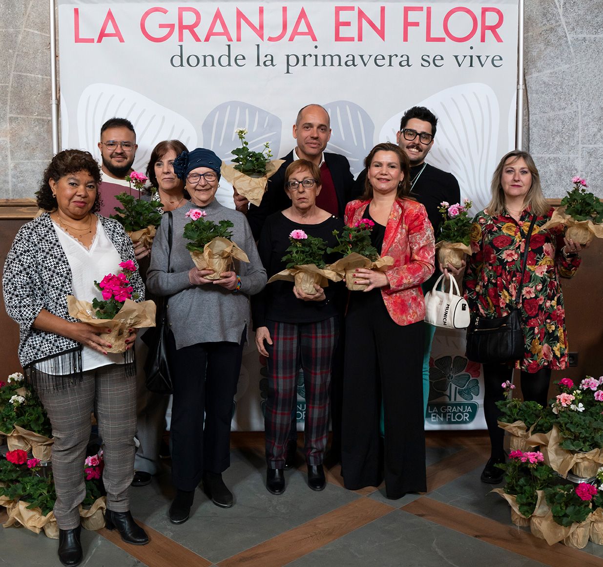Group of people holding geranium plants in front of La Granja en Flor spring festival promotional banner.