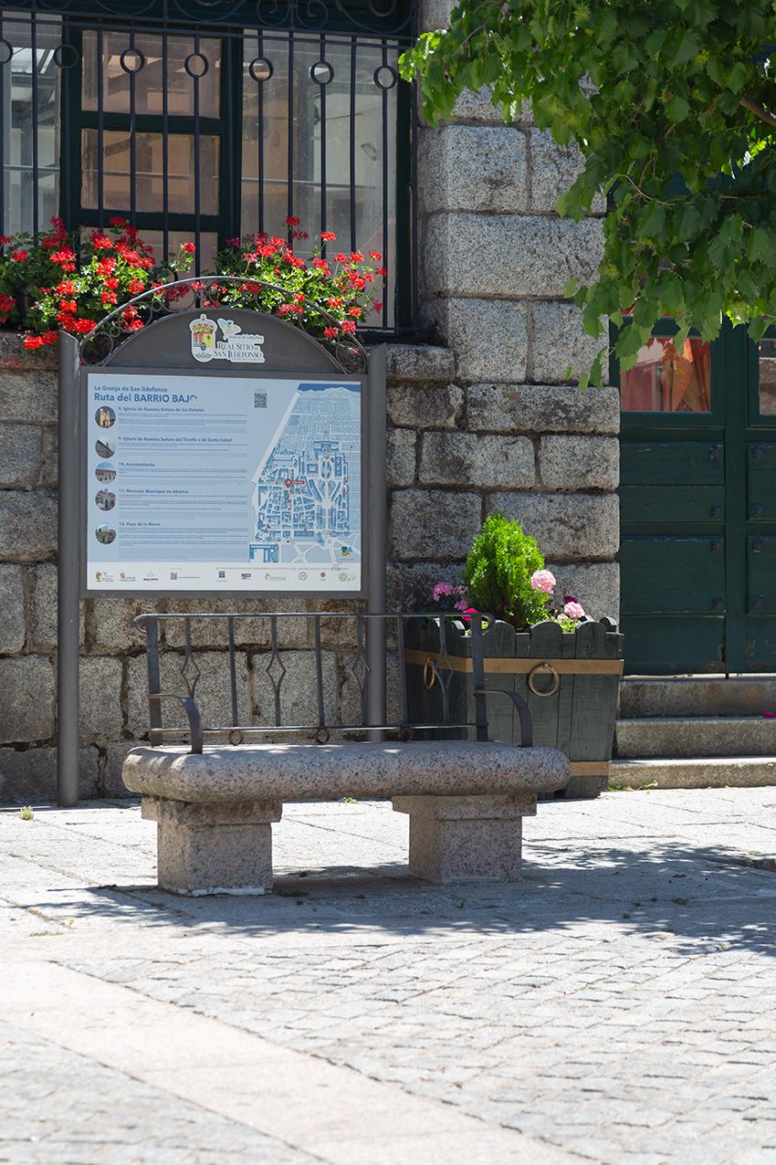 A stone bench sits beside an informational tourist map sign with flower planters near a granite building.