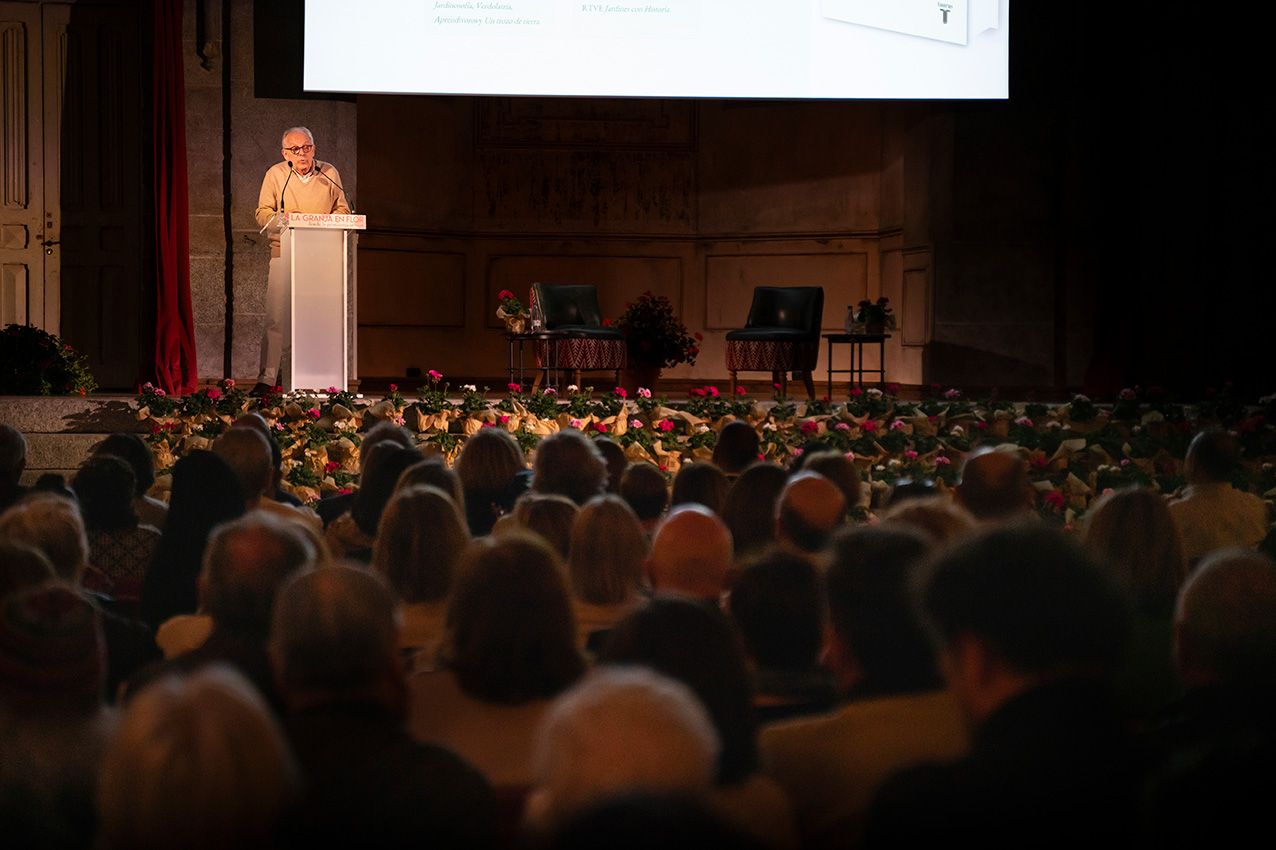 Speaker at podium addresses large audience in ornate hall decorated with flowers at a garden art conference.