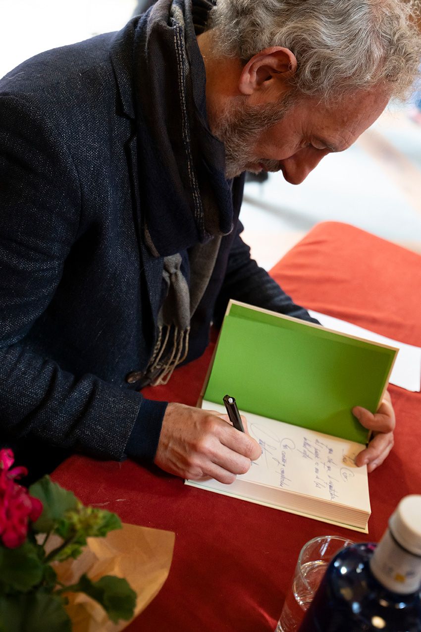 A bearded man with gray hair signs a green book at a red-draped table during a book signing event.
