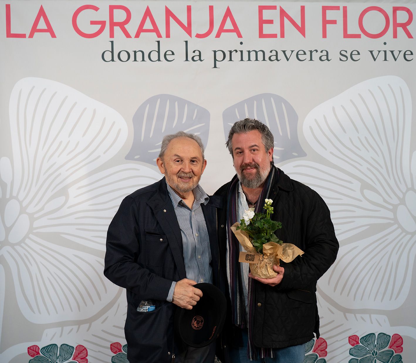 Two men posing together in front of La Granja en Flor spring festival banner.