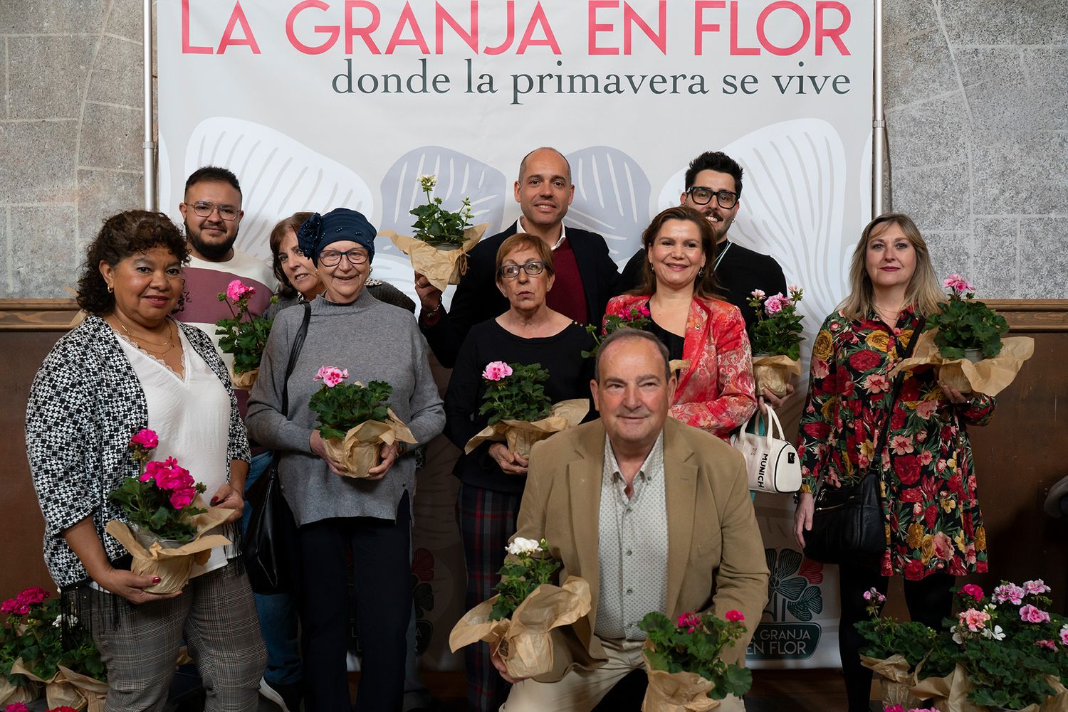 Group of smiling attendees posing with potted geraniums at La Granja en Flor donde la primavera se vive event.