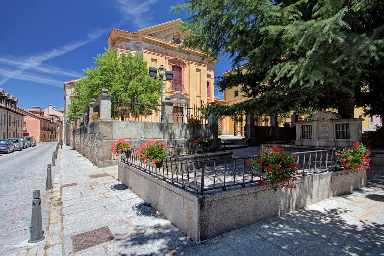 Historic European street with iron-fenced fountain, red geraniums, and grand yellow building surrounded by lush green trees.