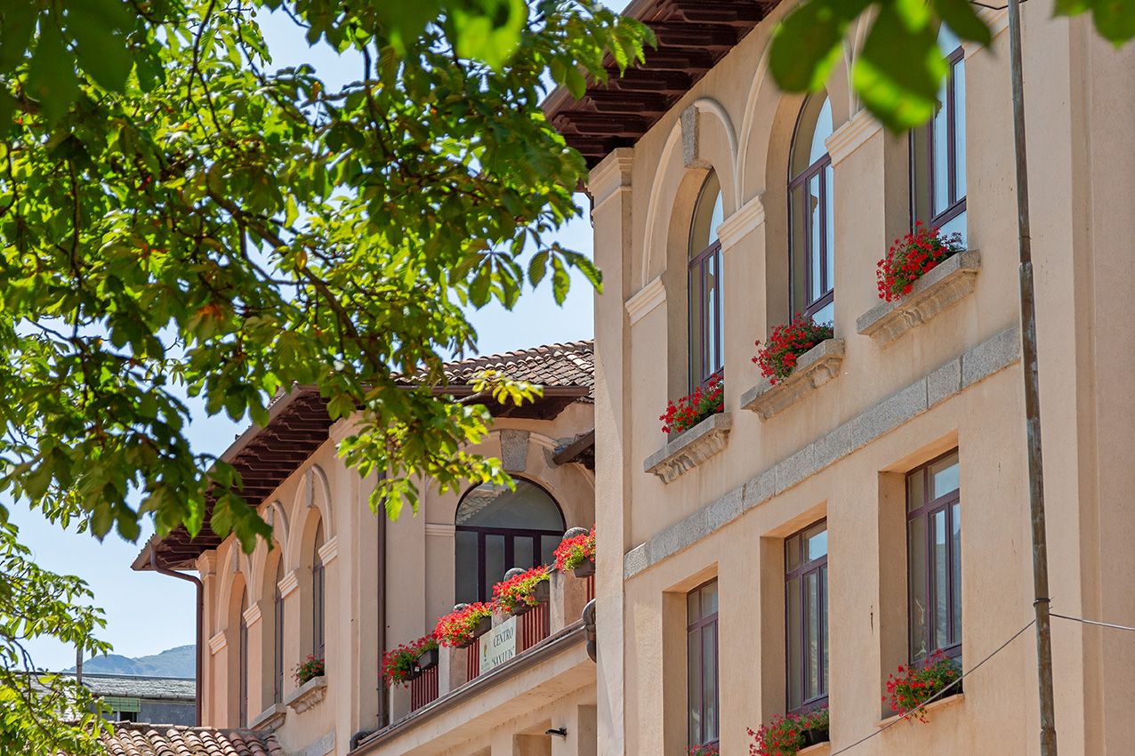 Historic European building facade with arched windows, red flower boxes, and lush green trees on a sunny day.