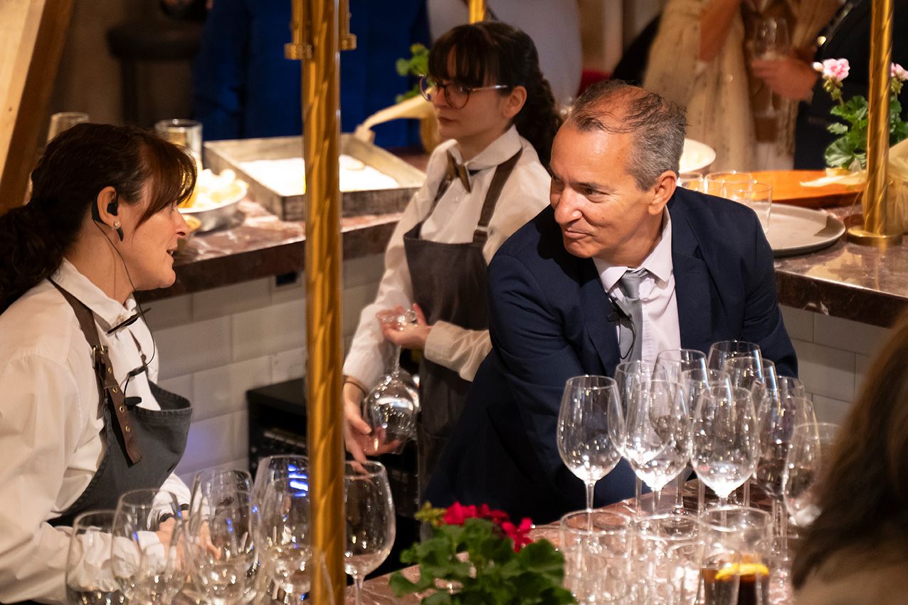 A man in a blue suit converses with a server at an elegant restaurant table set with wine glasses.