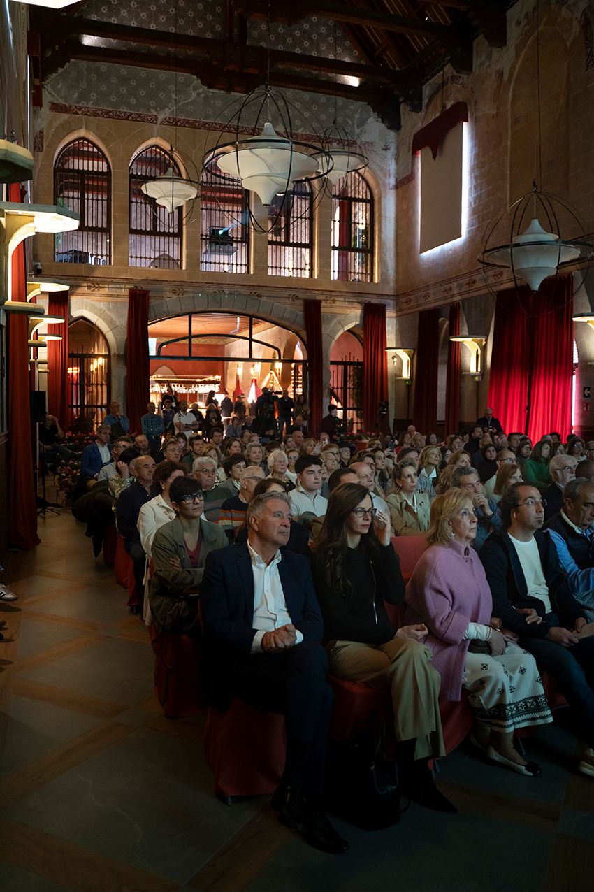 Large audience seated in historic ornate hall with arched windows and red curtains during a conference.