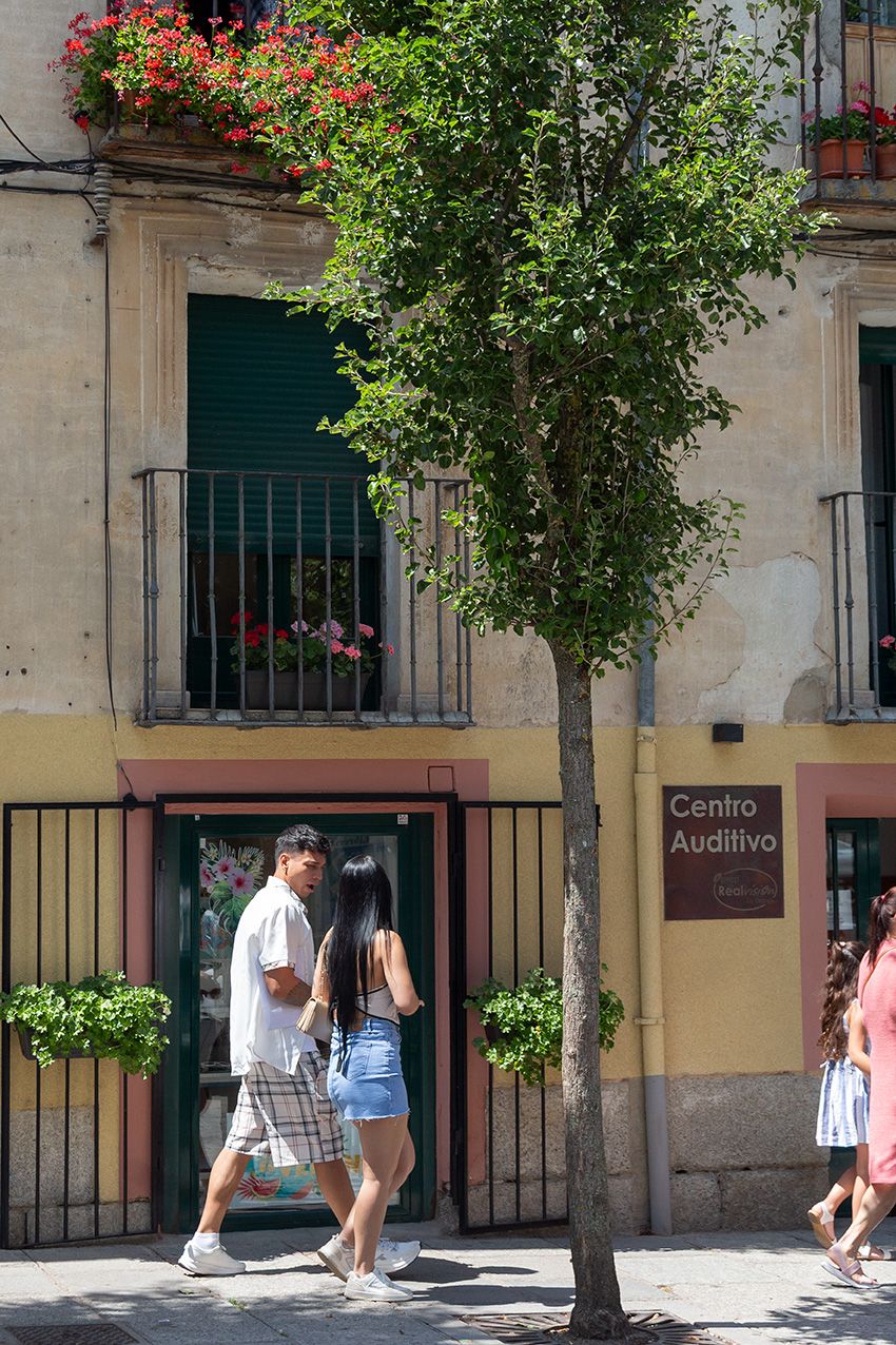 Couple walking past a Centro Auditivo shop on a sunny European street with a tree and flower-adorned balconies.