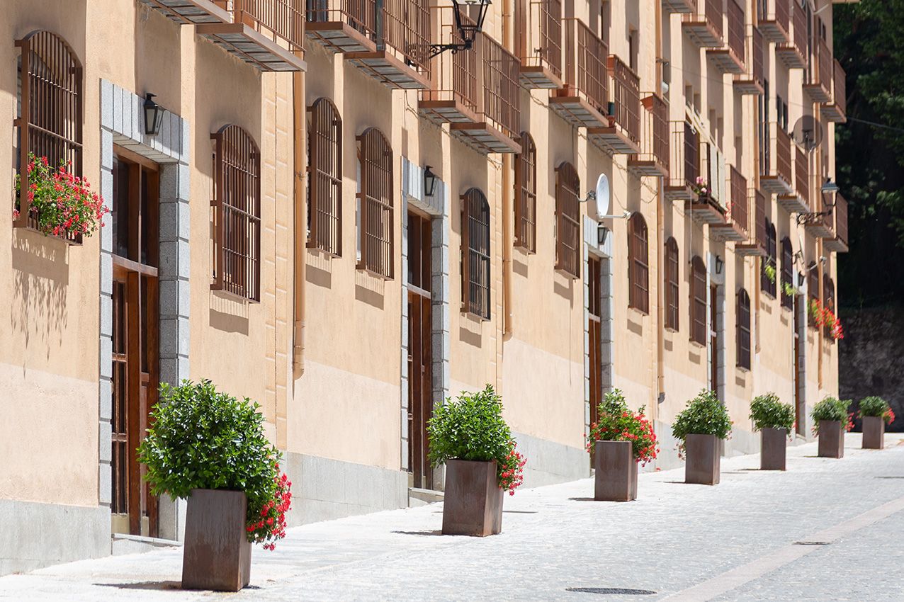 A charming European street lined with potted plants and flower boxes adorning a beige building facade with wooden doors.