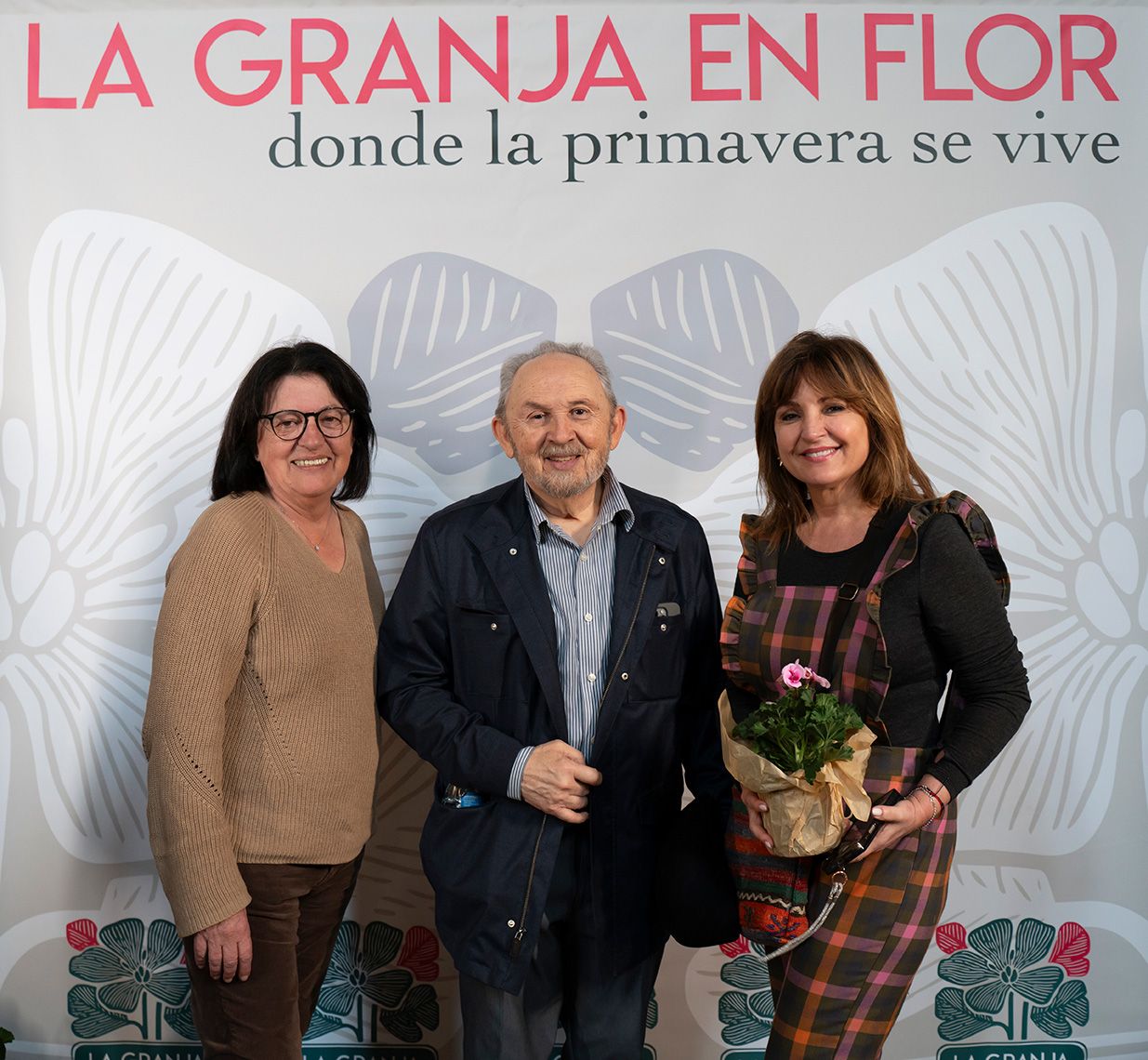 Three people posing at La Granja en Flor spring event backdrop, woman holding potted pink flower.