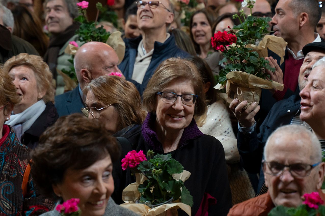 Smiling woman with glasses holds pink geraniums among a cheerful crowd at an indoor flower event.