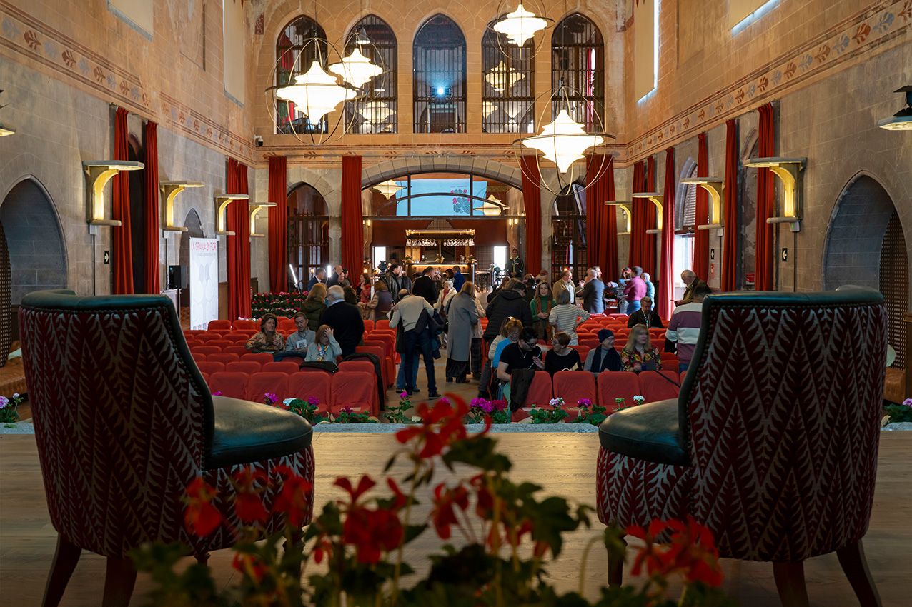 Ornate grand hall with red carpets, arched windows, and chandeliers filled with visitors.