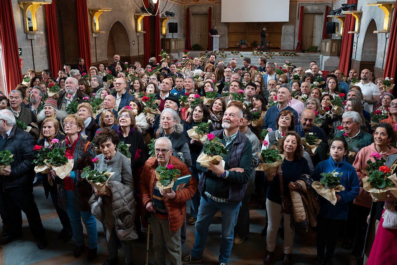 Large group of people holding potted geraniums inside an ornate hall at La Granja en Flor flower festival.