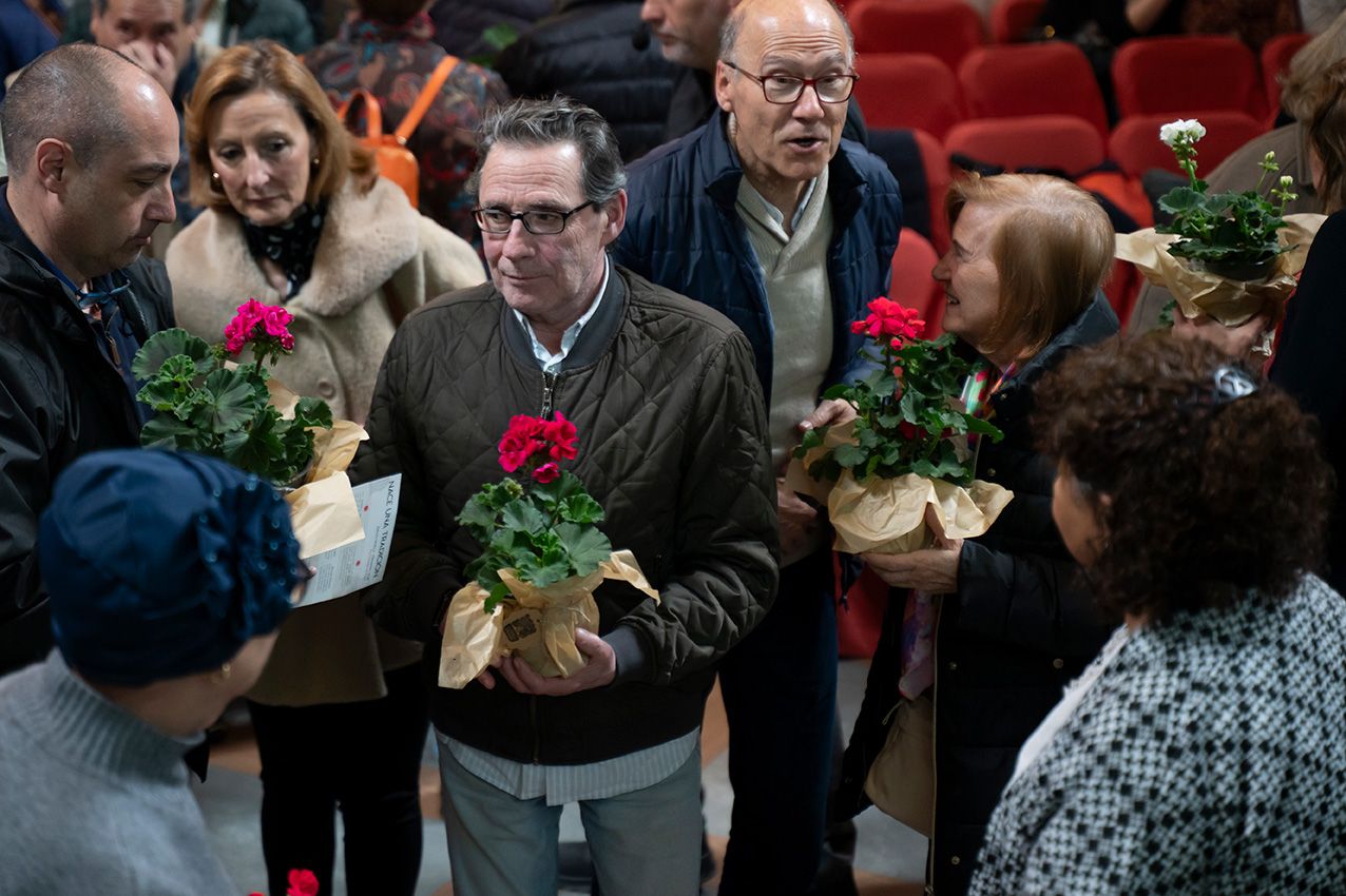 Group of people holding potted geraniums wrapped in gold paper at indoor event.