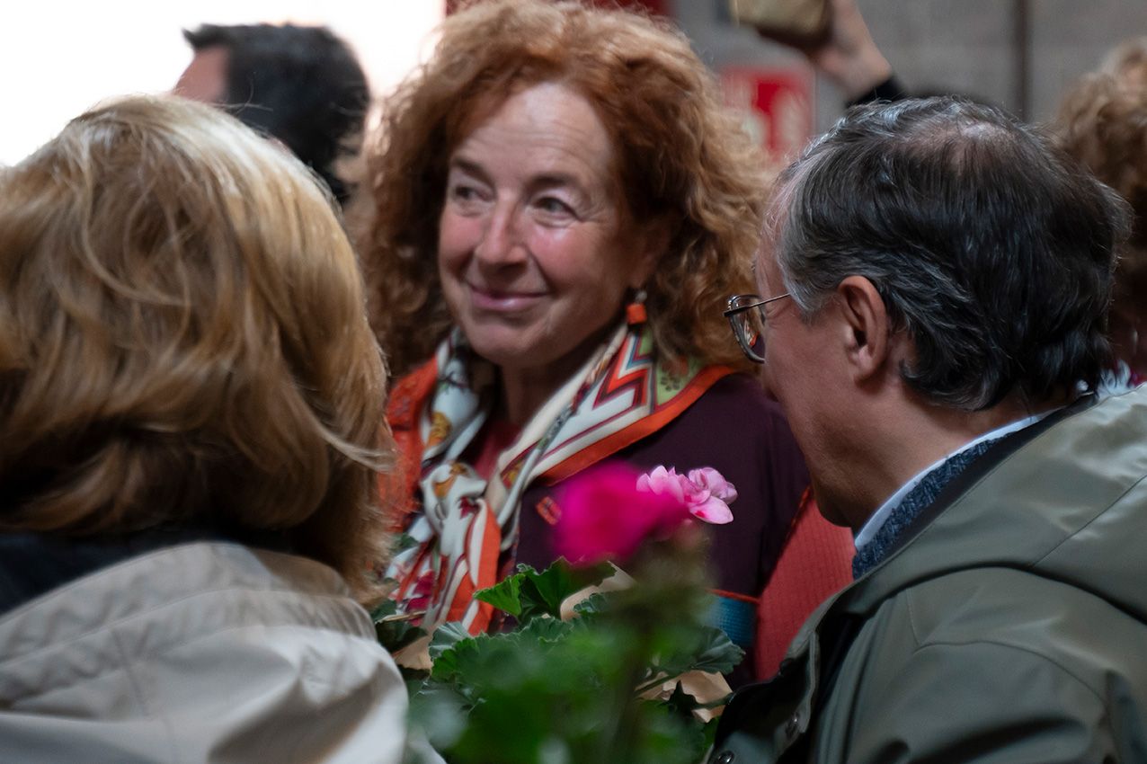 Smiling red-haired woman with colorful scarf holding pink flowers among a crowd at a social gathering.