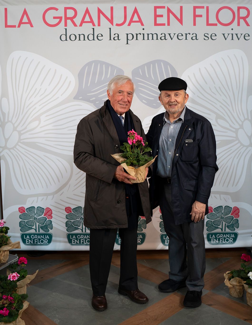 Two elderly men posing together at La Granja en Flor event, one holding a pink potted plant.