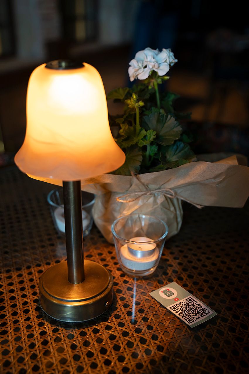 Glowing table lamp beside a candle, white flowers, and QR code card on wicker surface in dimly lit restaurant.