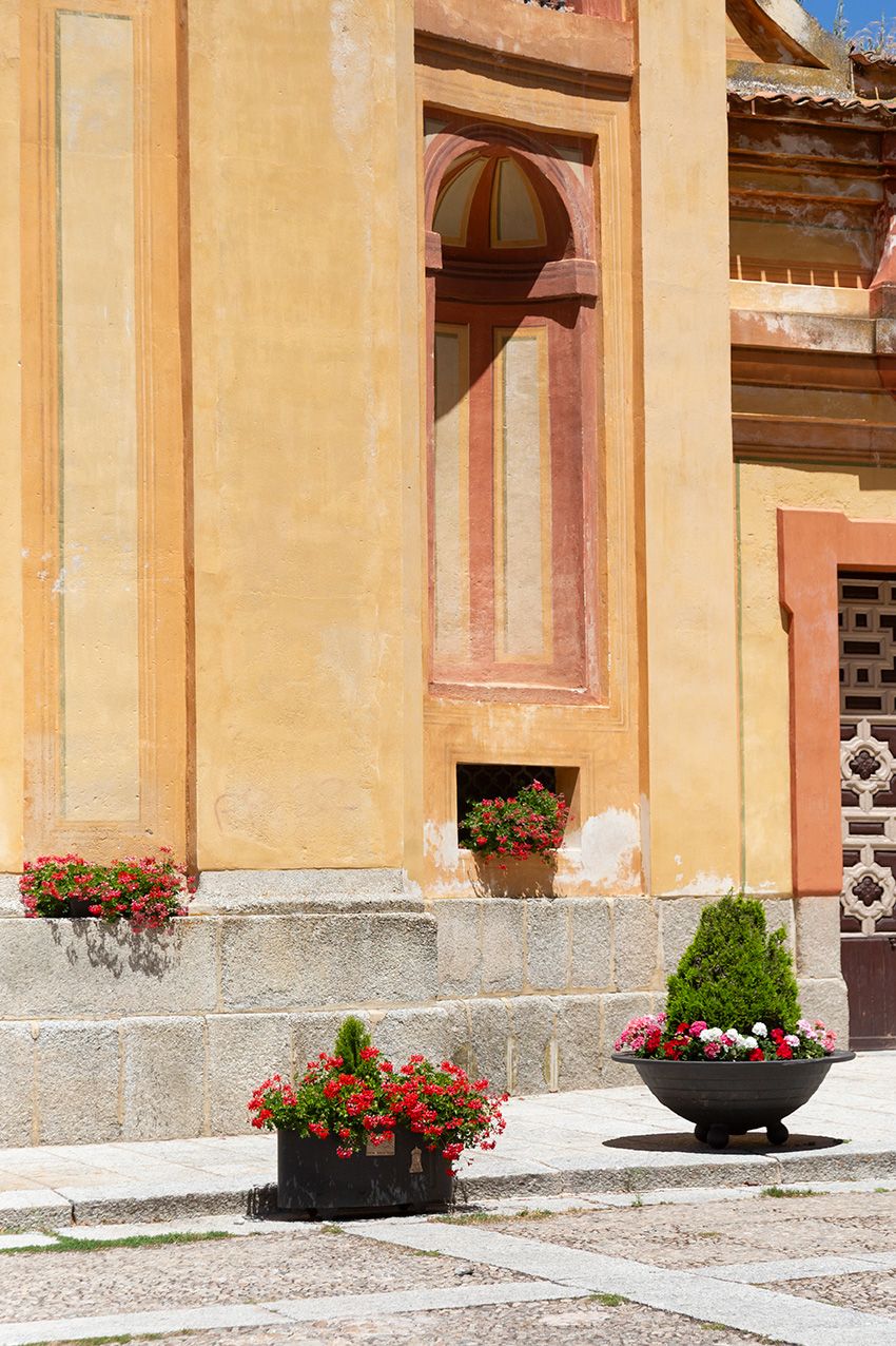 Historic yellow building facade with arched windows, stone steps, and decorative flower pots with red blooms in a sunny courtyard.