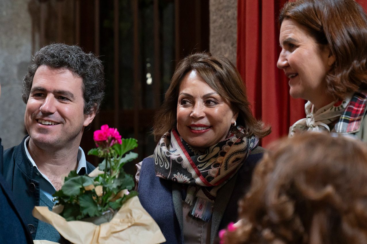 Three people smile together at outdoor event, woman in center holds wrapped pink rose bouquet.