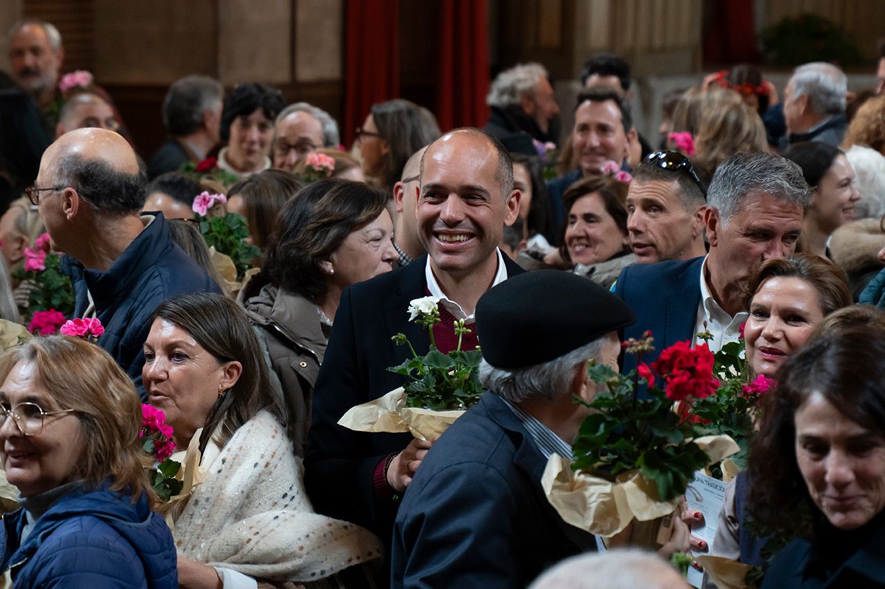 Smiling man holds colorful geranium bouquets surrounded by a crowd at an indoor flower ceremony.
