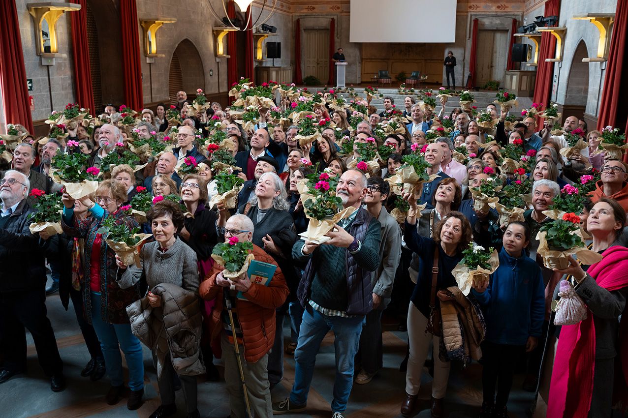 Crowd of attendees holding potted geraniums in a grand decorated hall at La Granja en Flor spring festival.