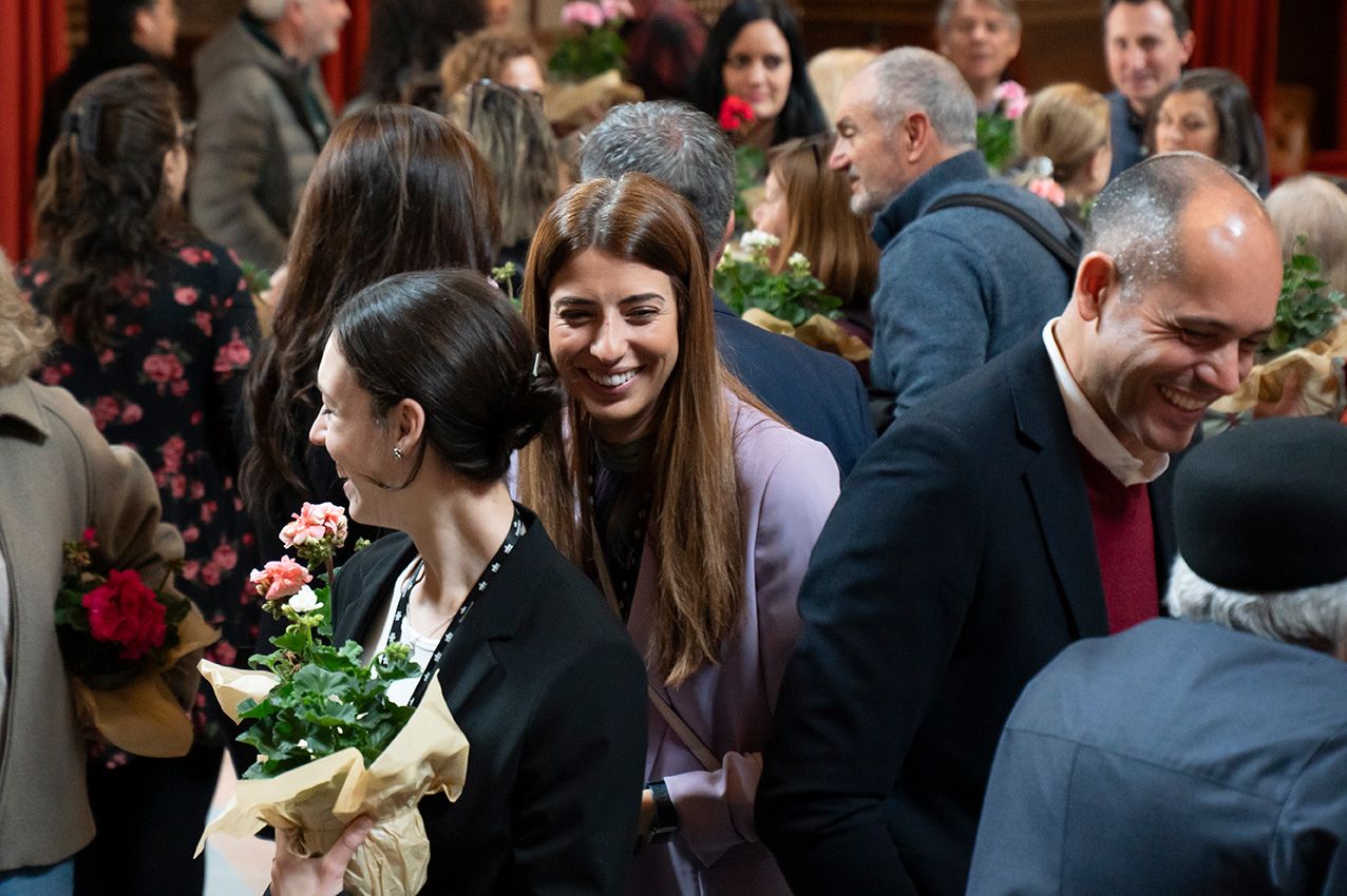 Young smiling woman holding a pink geranium plant among a crowd at an indoor social gathering.
