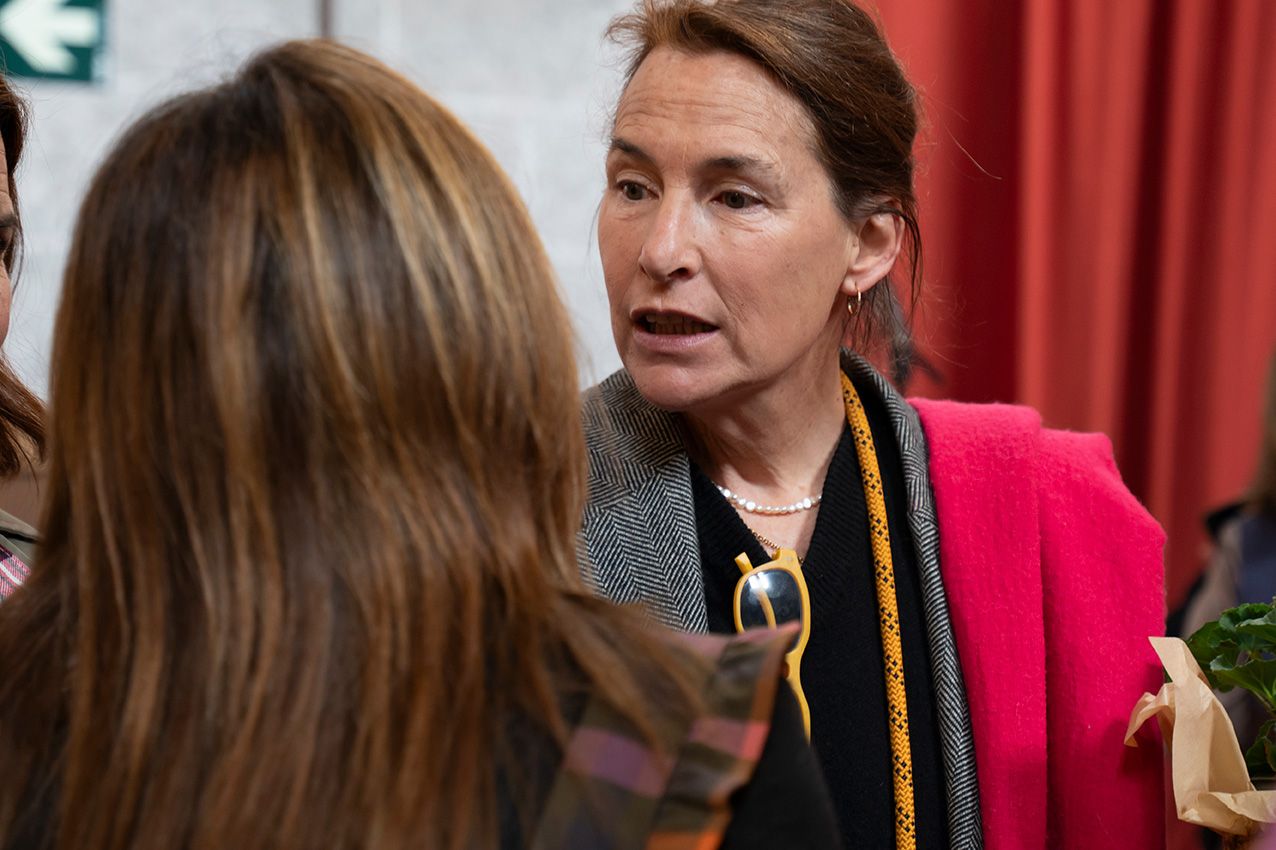 Woman in red blazer with pearl necklace engaged in conversation at an indoor event.