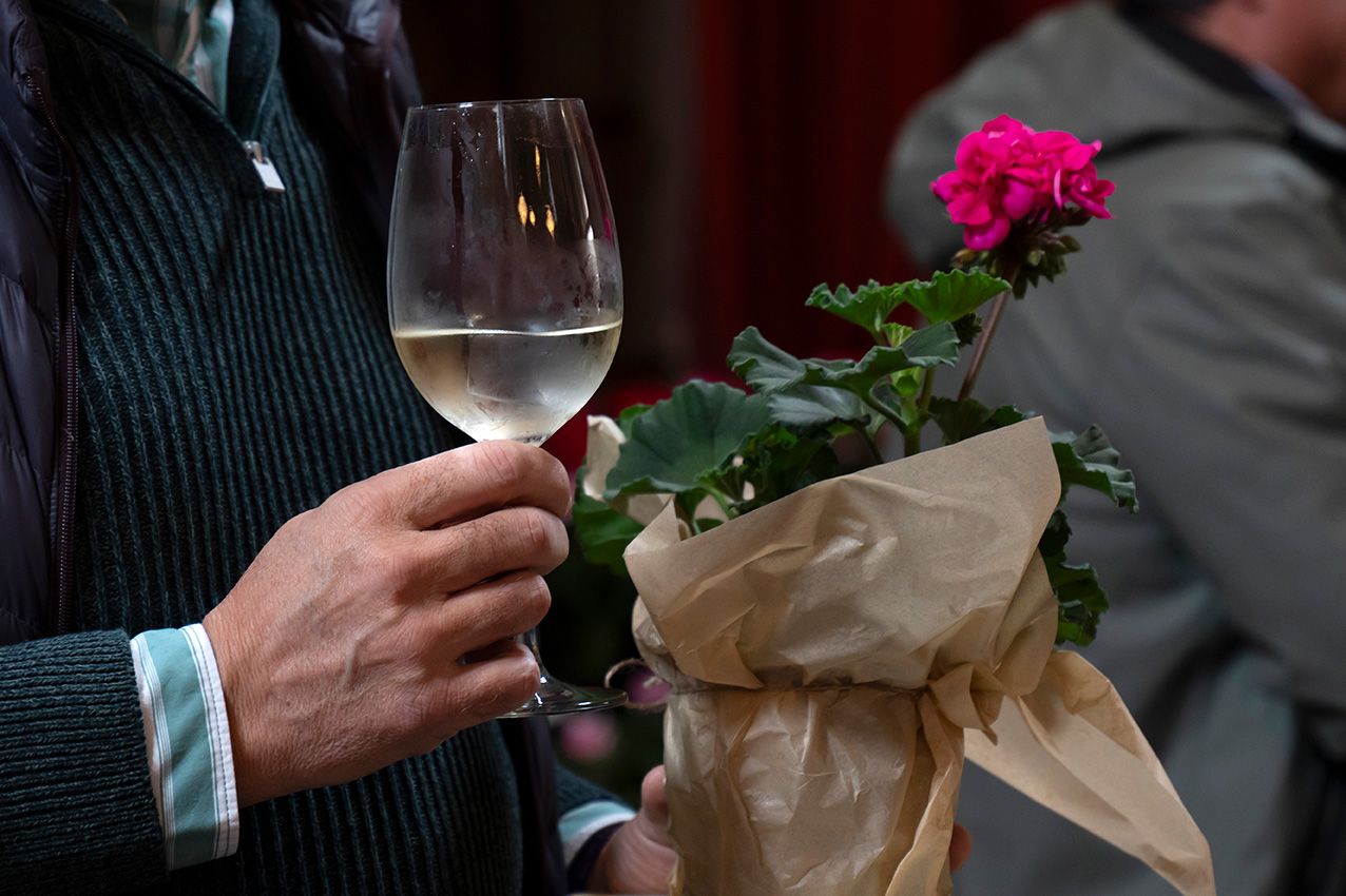 Person holds white wine glass and wrapped pink geranium plant at outdoor spring garden event.