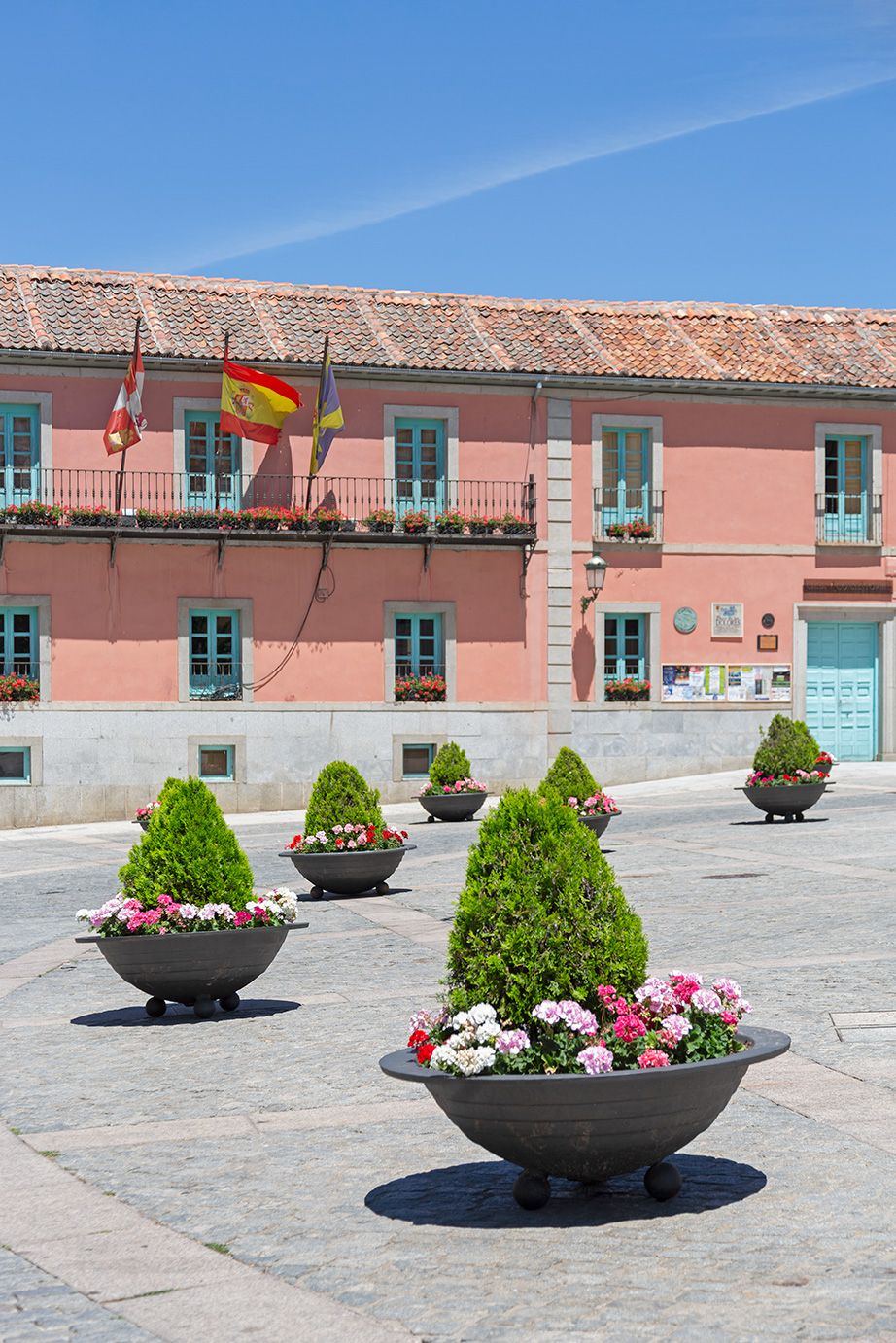 Pink building with teal shutters and Spanish flags, flanked by ornamental planters with topiaries and flowers in a sunny plaza.