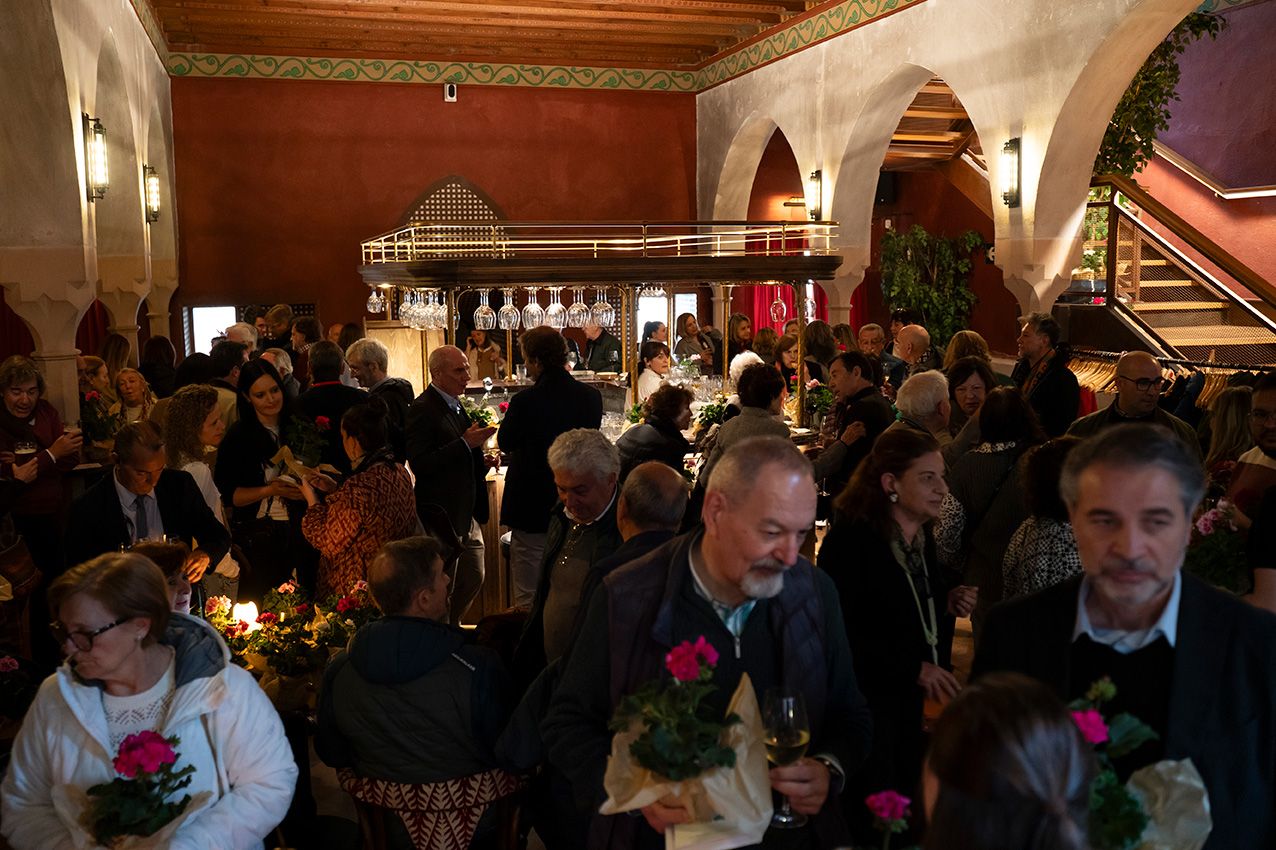 A large crowd mingles at a festive indoor event in a warmly lit historic venue with pink flowers.