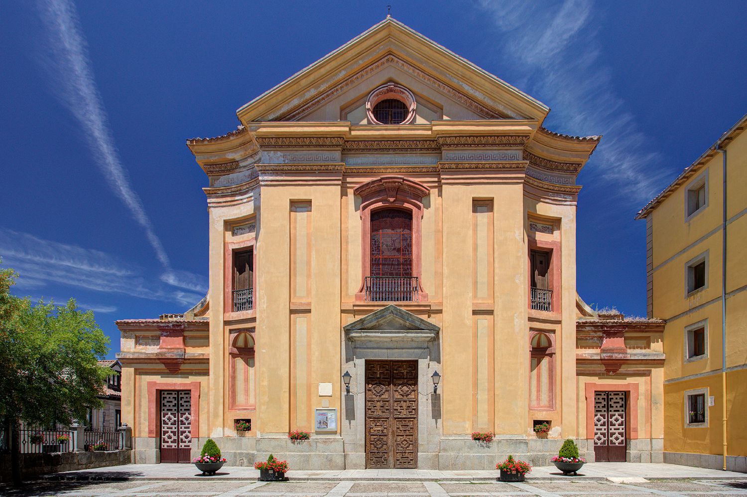 Baroque Italian church facade with peach stucco walls, ornate details, wooden doors, and flower pots under a blue sky.