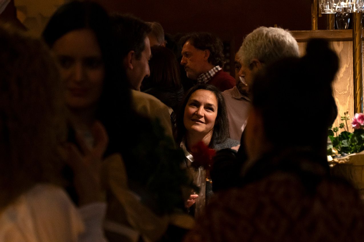 A smiling woman holds a wine glass while engaging in conversation at a dimly lit social event.