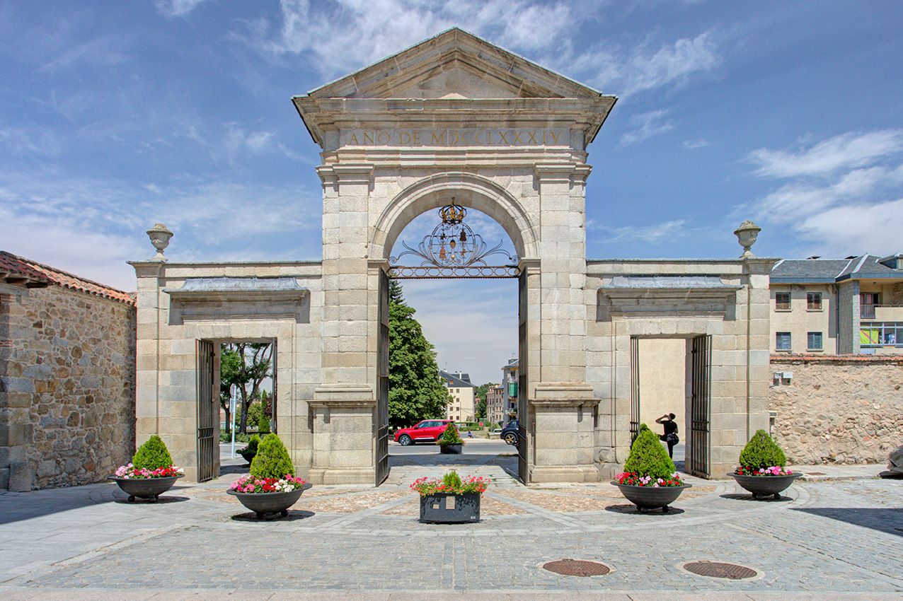 Grand stone archway entrance gate with ornate pediment, flanked by decorative planters and manicured trees under blue sky.
