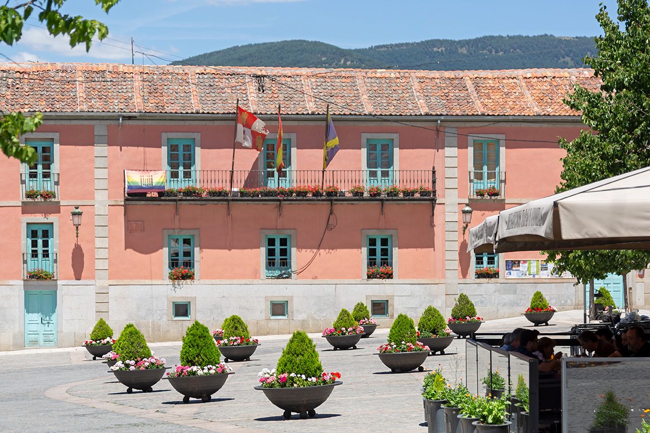 Pink Spanish town hall with teal shutters and flags, surrounded by decorative planters with topiaries in a sunlit square.
