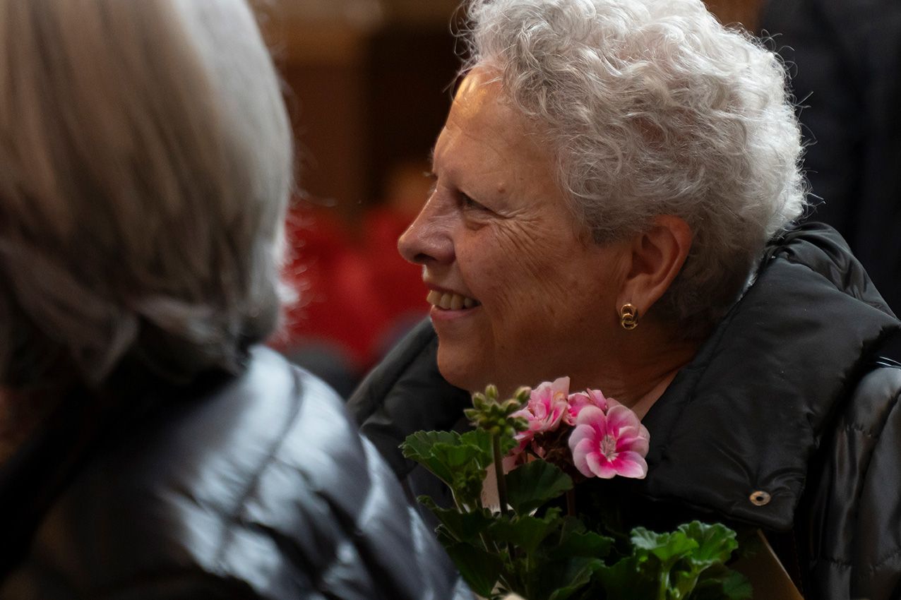 Smiling elderly woman with white hair holding a pink geranium flower bouquet at an outdoor event.